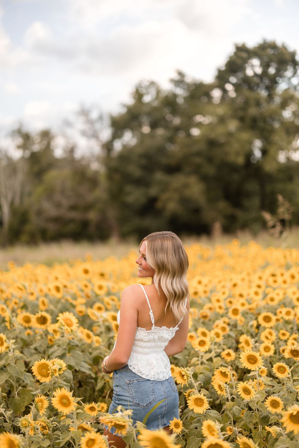 A woman in a white top sits among a vibrant field of blooming yellow sunflowers on a sunny summer day.