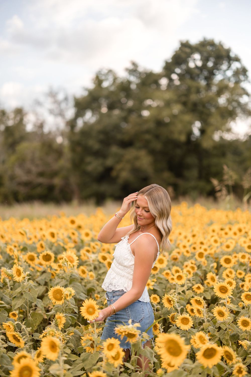Person in white tank top and denim shorts poses among vibrant yellow sunflowers in a summer field.