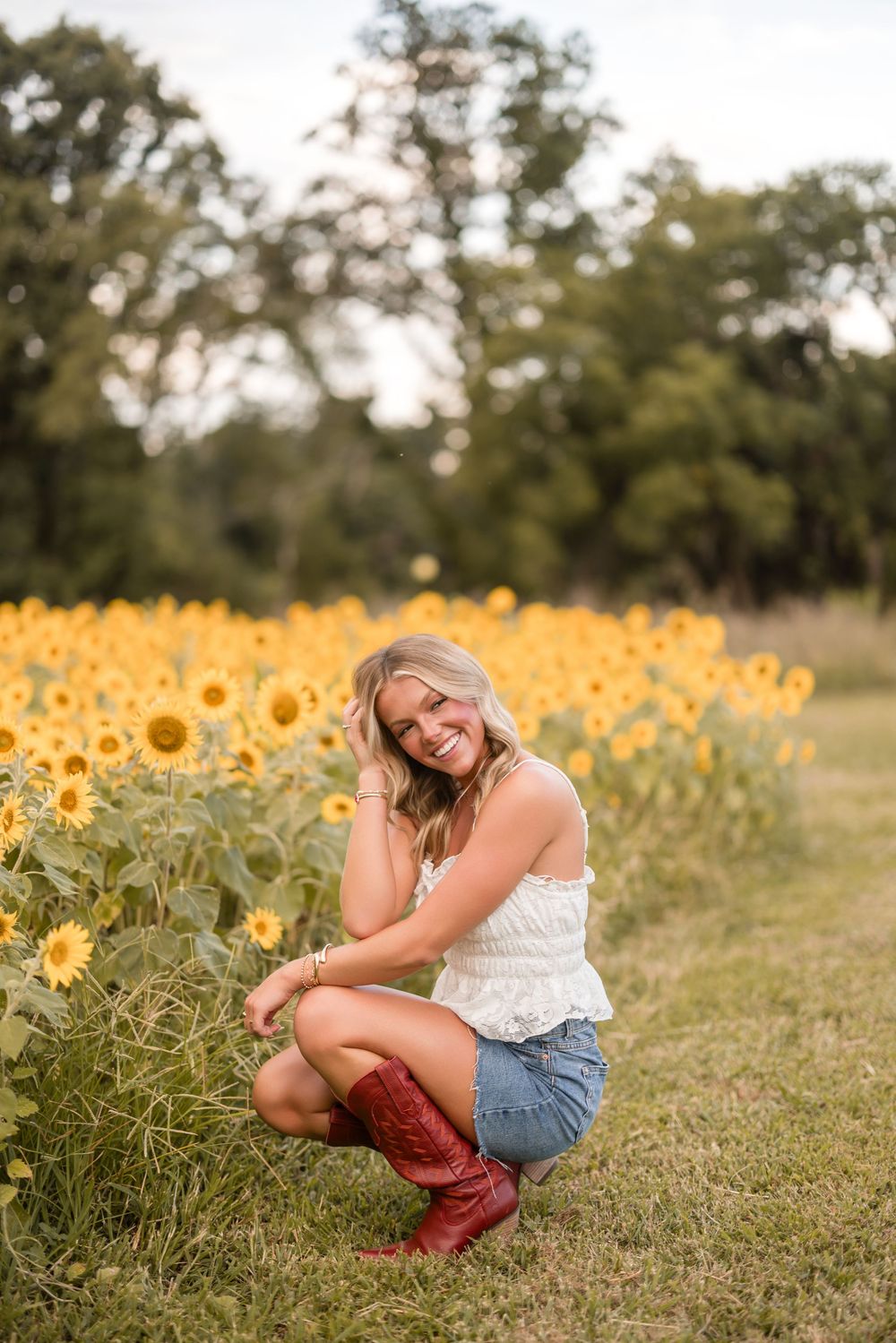 A figure sits among blooming sunflower fields in a summer landscape wearing a white top and denim shorts.