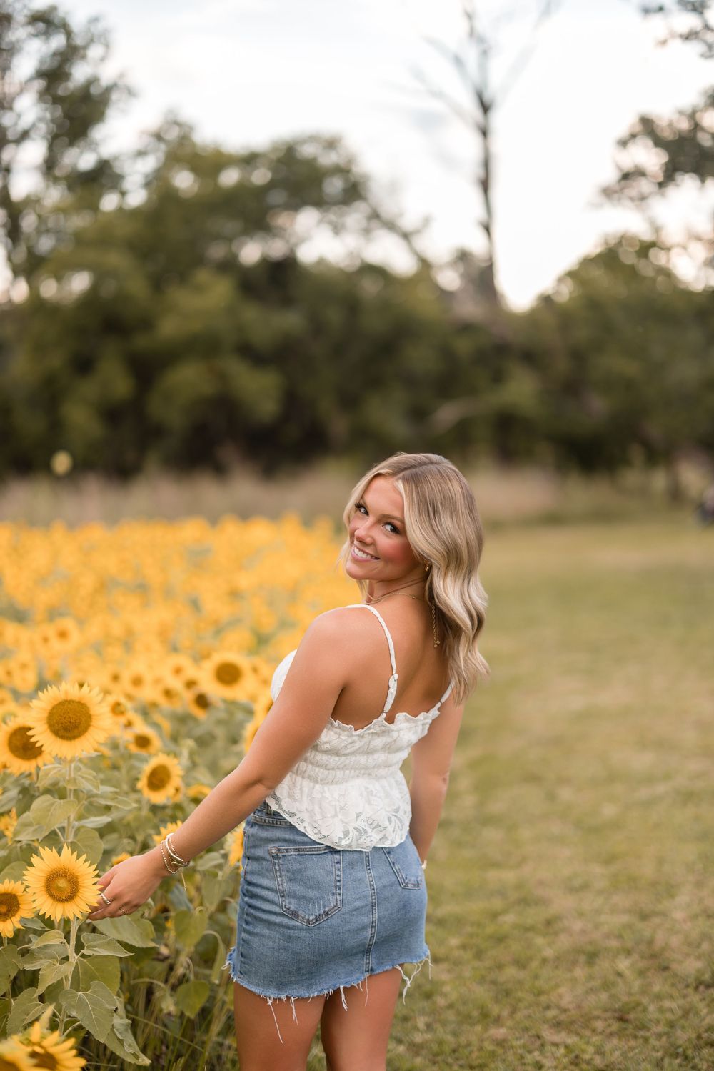 A young woman in a white top and denim skirt poses by a vibrant field of blooming sunflowers at sunset.