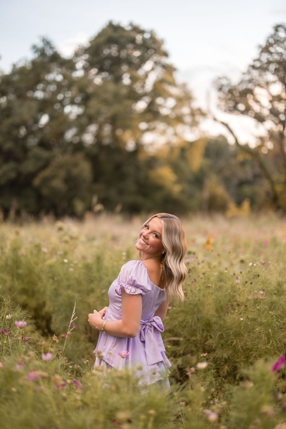 A woman in a lavender dress enjoys a summer evening in a wildflower meadow while golden sunlight filters through trees.