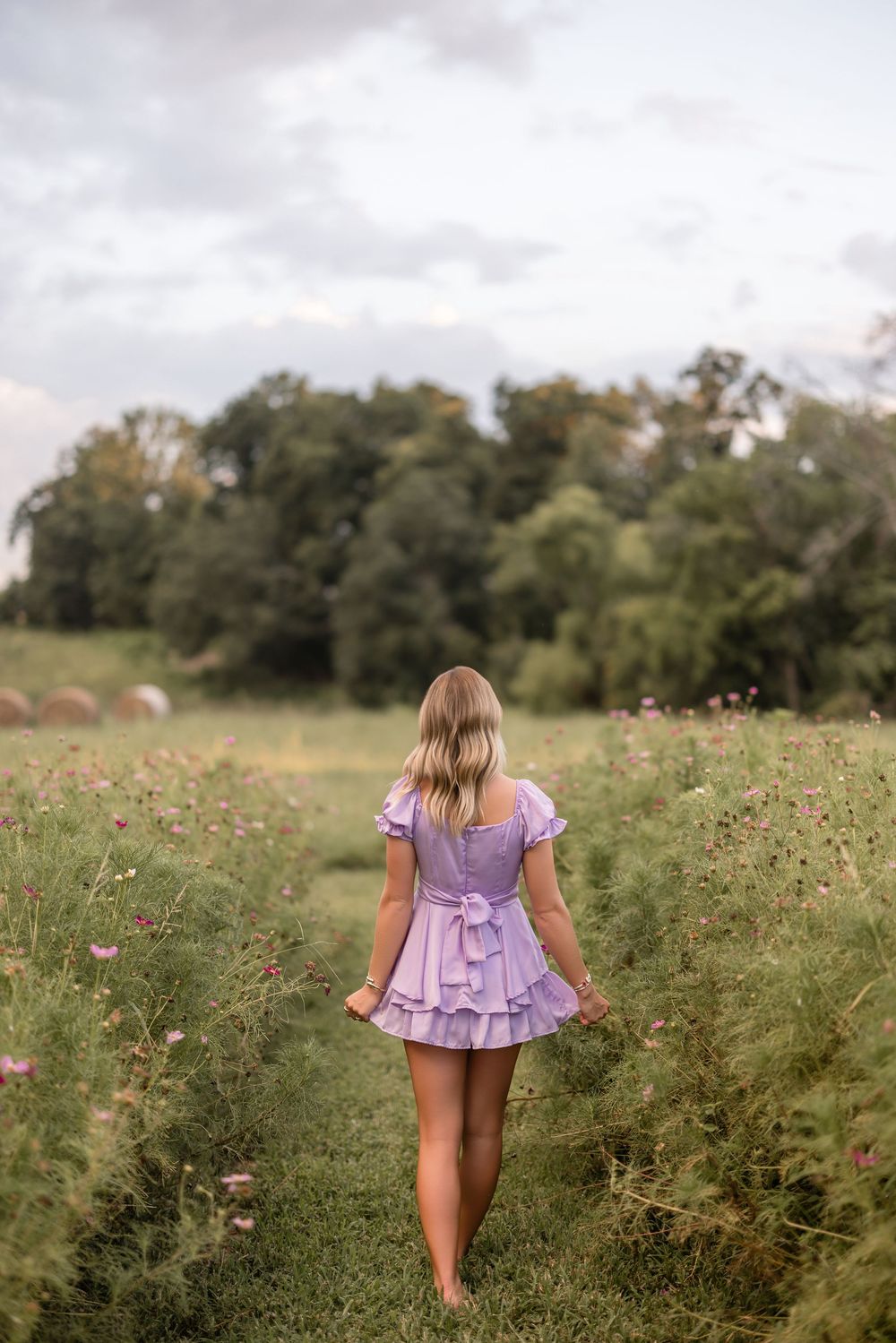 A person in a lilac dress walks through a wildflower meadow on a cloudy summer day.