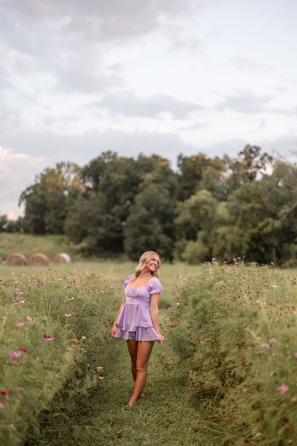 Figure in a purple dress twirls joyfully in a wildflower meadow with hay bales and trees visible in the background.