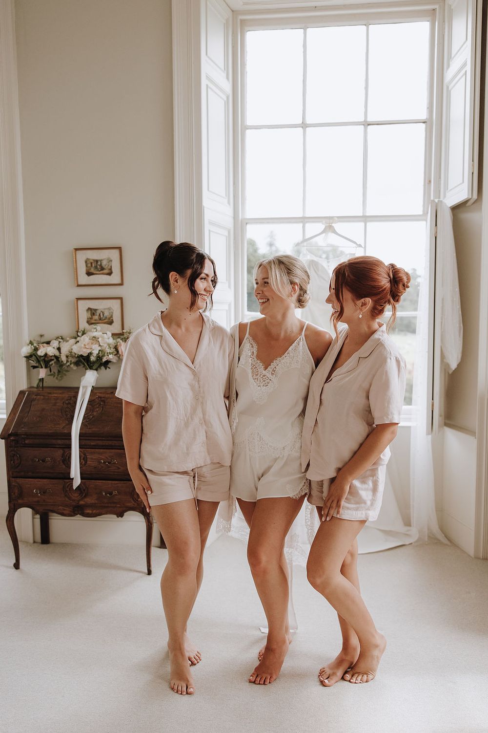 Three people in matching white satin pajama sets stand together barefoot by a bright window in an elegant room.