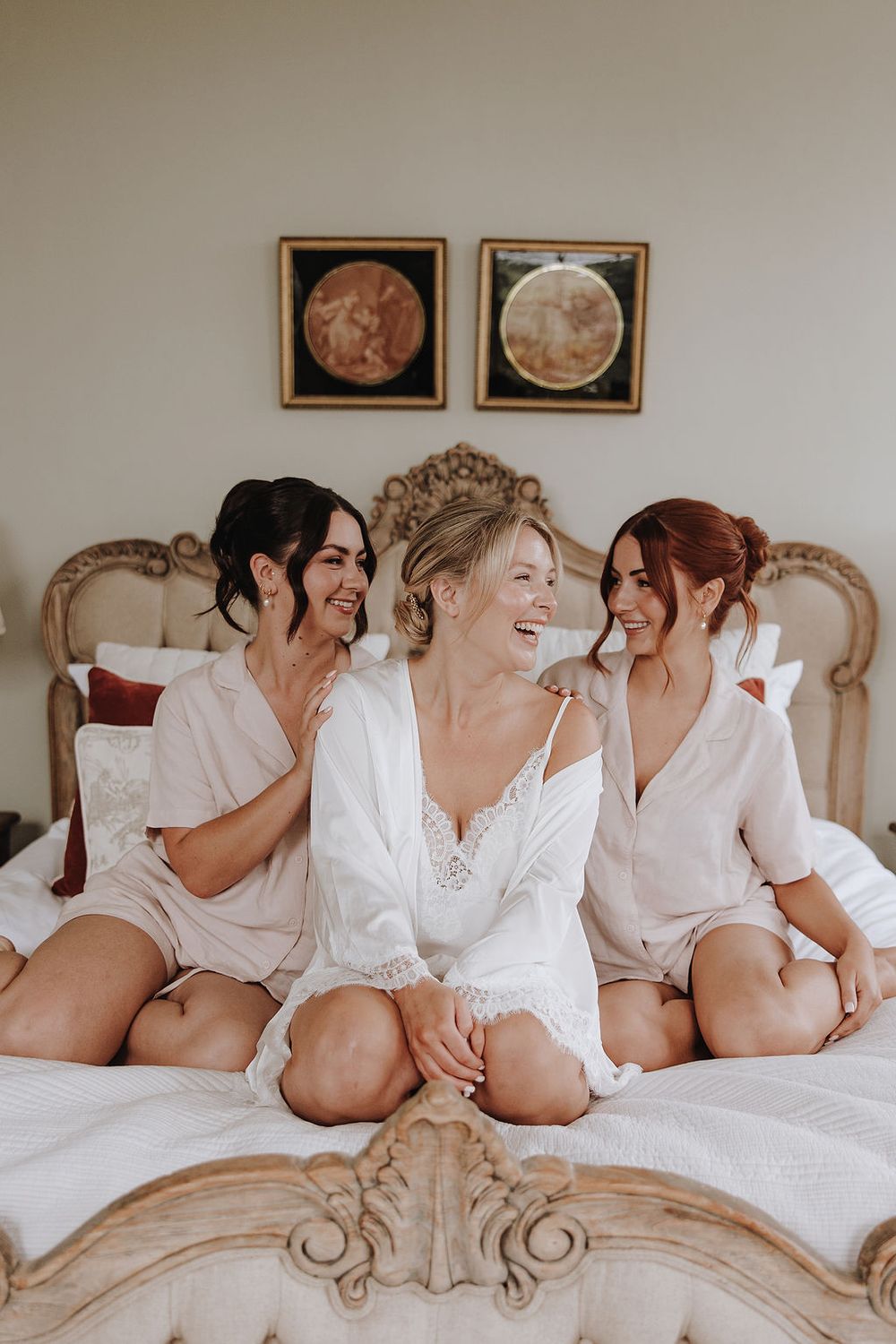 Three women in white robes share joyful laughter while sitting together on an ornate vintage bed.