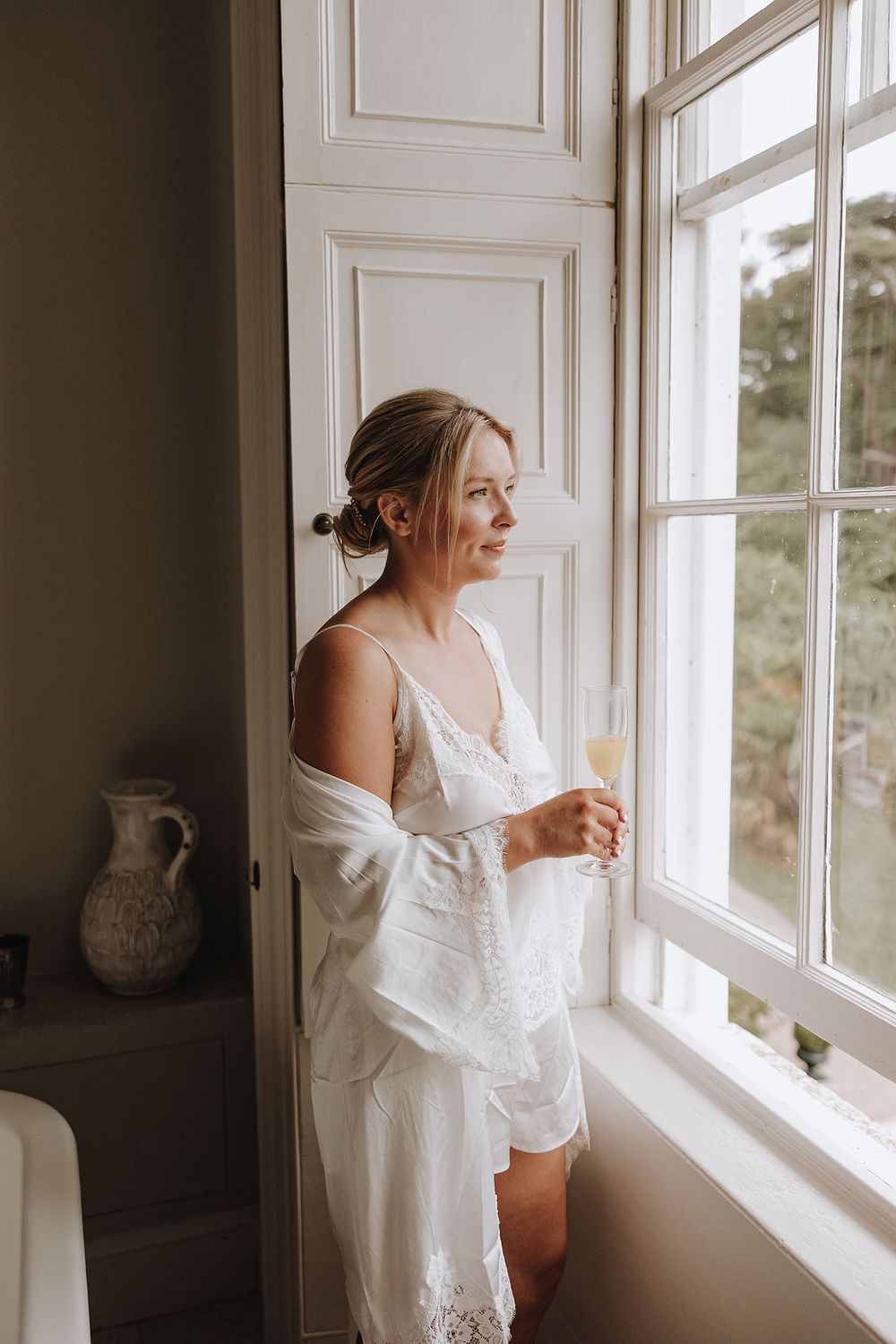 A figure in a flowing white dress gazes thoughtfully through a bright window in an elegant interior space.