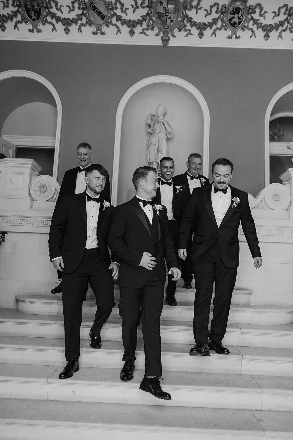 Group of groomsmen in black tuxedos walk down elegant white stone steps in sequence shot.