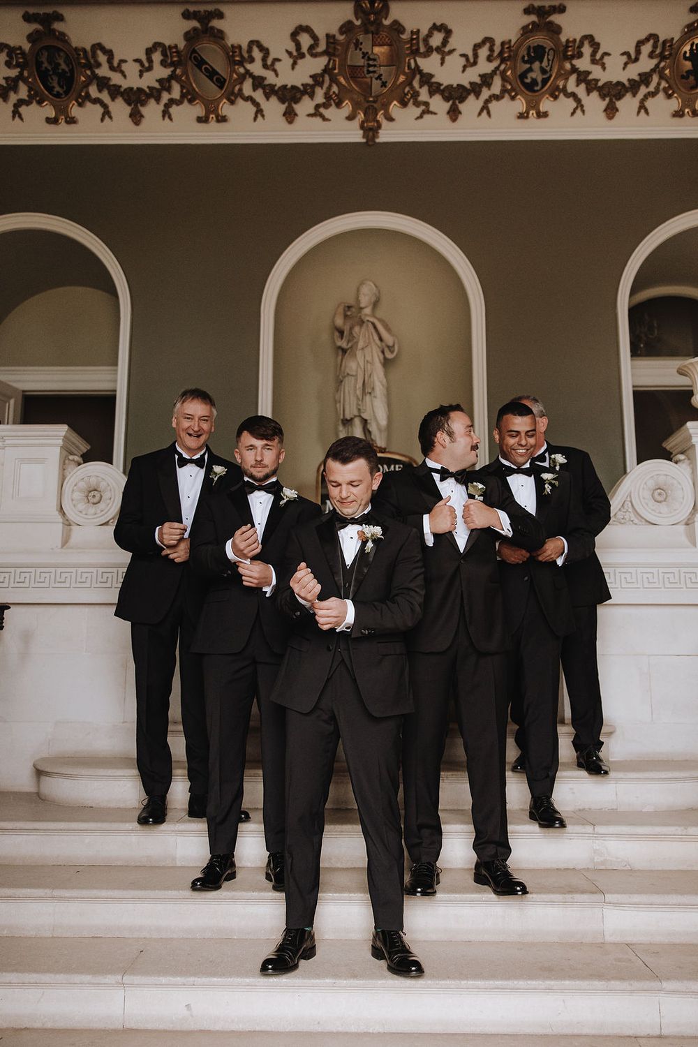 Groomsmen in black tuxedos and bowties pose elegantly on white marble steps with an ornate arch and statue in background.