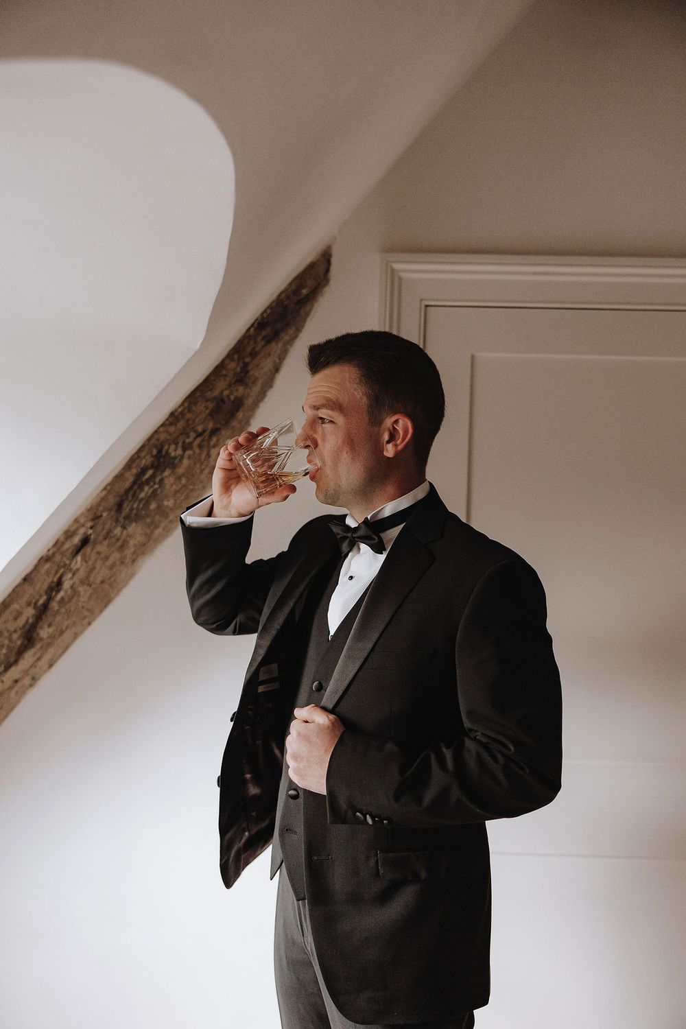 Someone wearing a formal black tuxedo and bow tie takes a drink near a white wall with exposed wooden beams.
