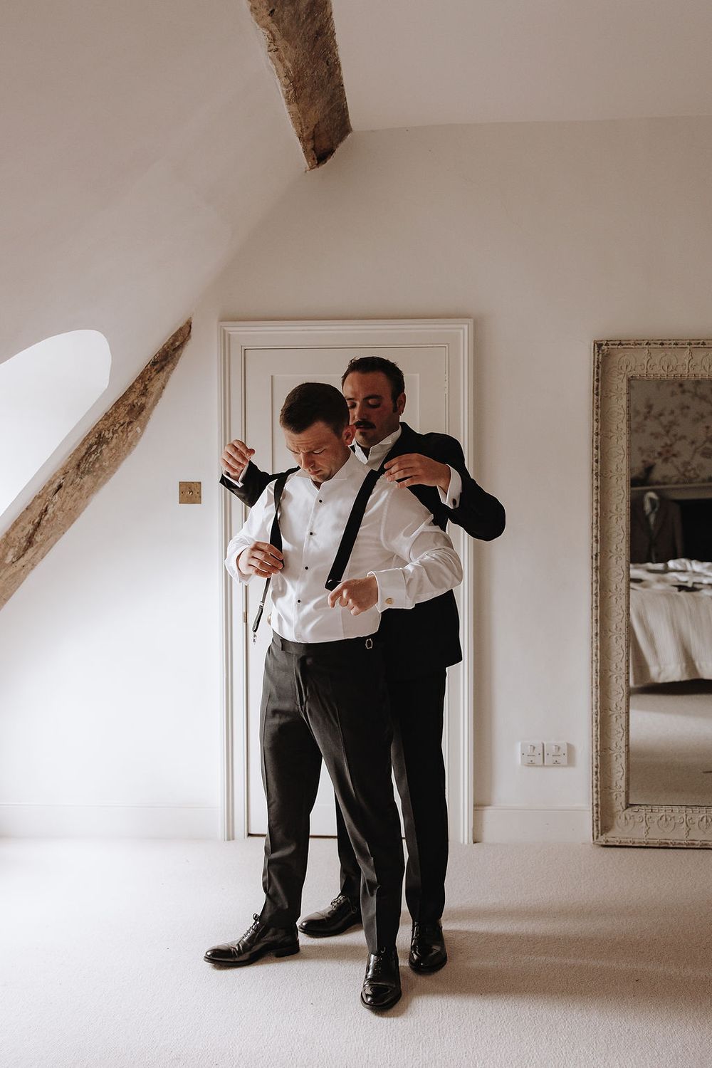 Two people in formal attire sharing a candid moment while getting ready in a bright attic room with exposed wooden beams.