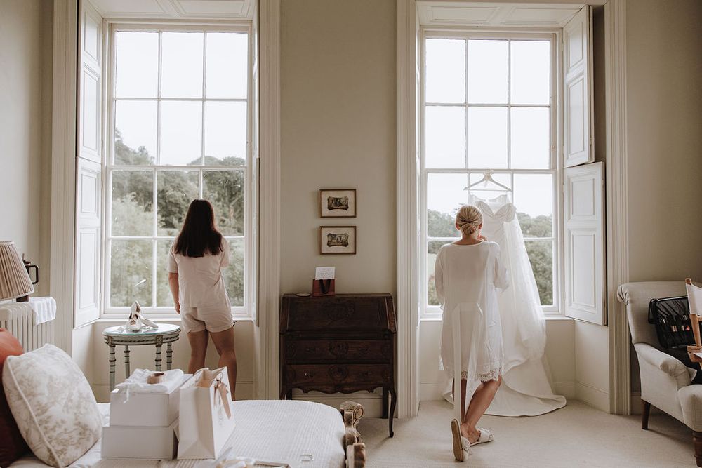 Two people in white dresses stand by tall windows in an elegant bright room with vintage furniture and decor.