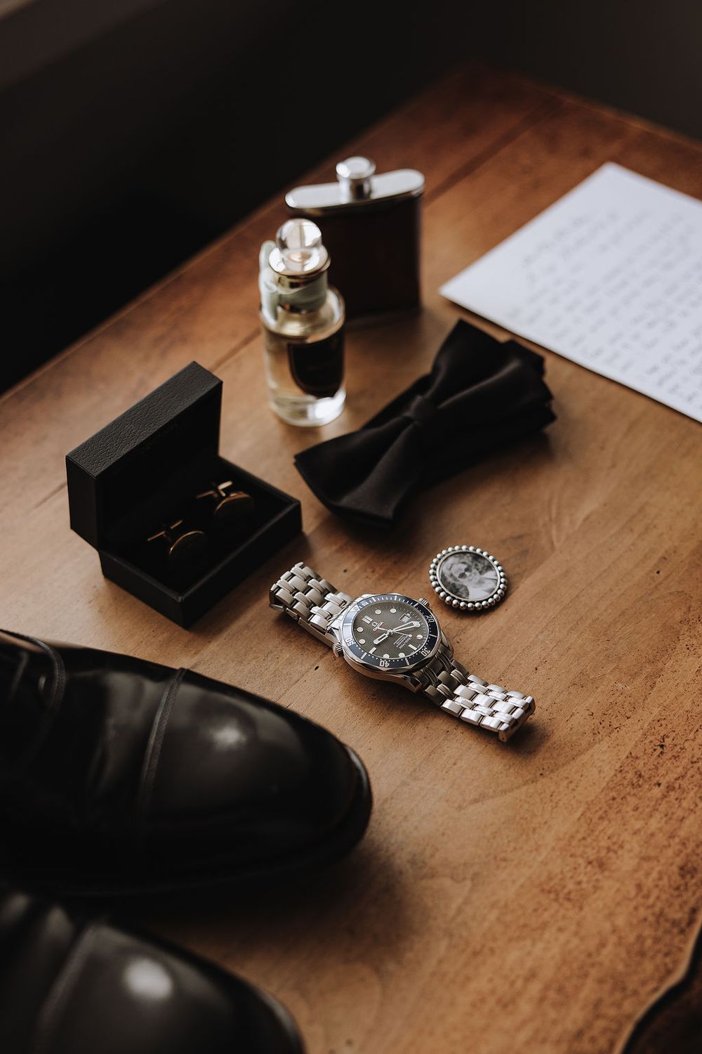 Men's formal accessories including a wristwatch, bow tie, ring box, and dress shoes arranged on wooden surface with perfume.
