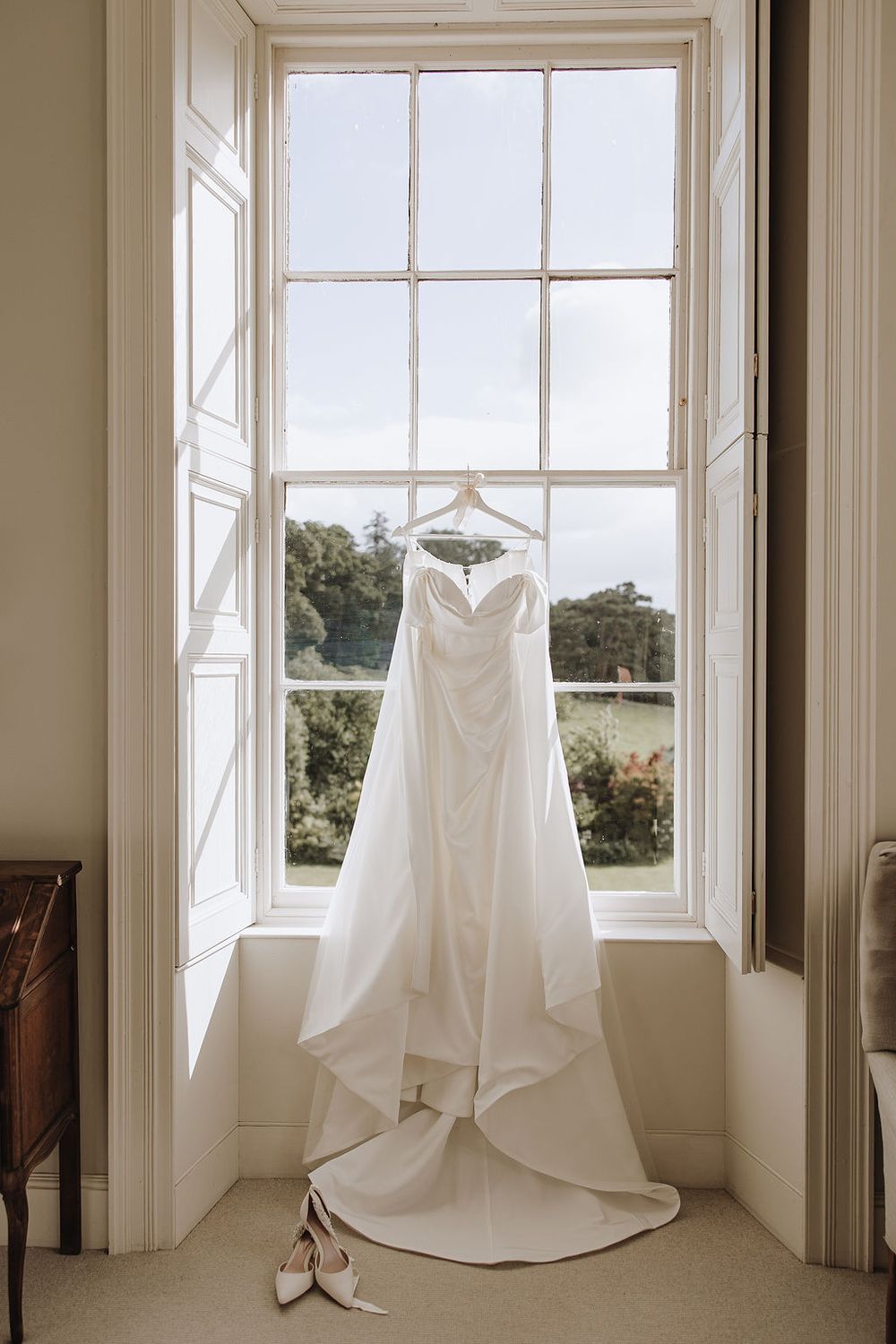 A white wedding dress and bridal shoes hanging in front of a tall window with countryside views.