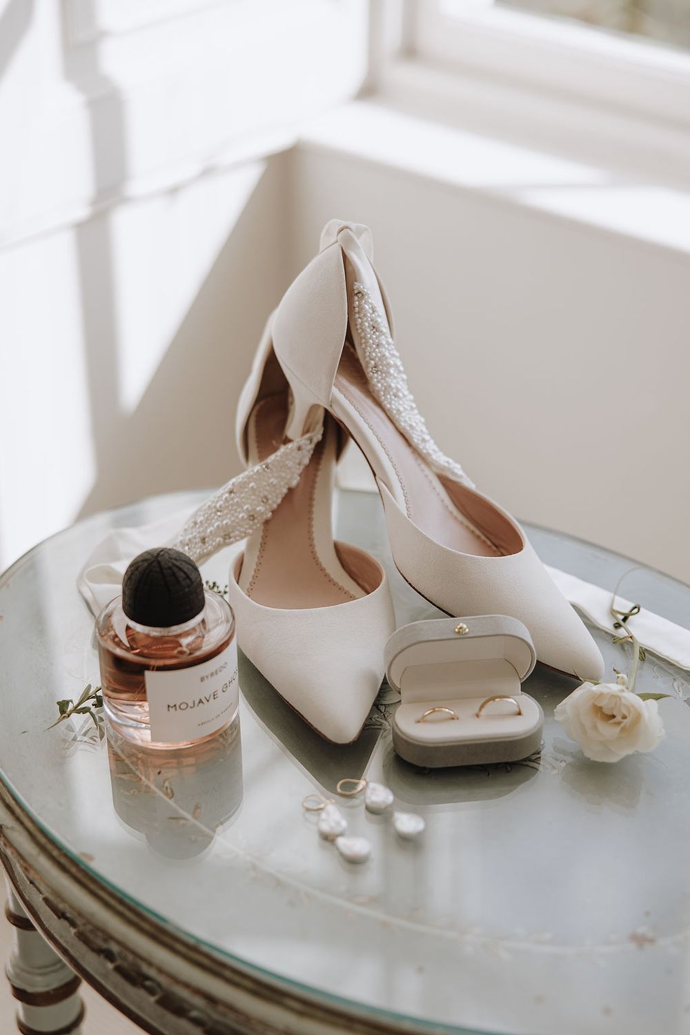 Elegant white wedding heels displayed with perfume bottle, ring box, and pearls on vintage mirrored table in soft light.