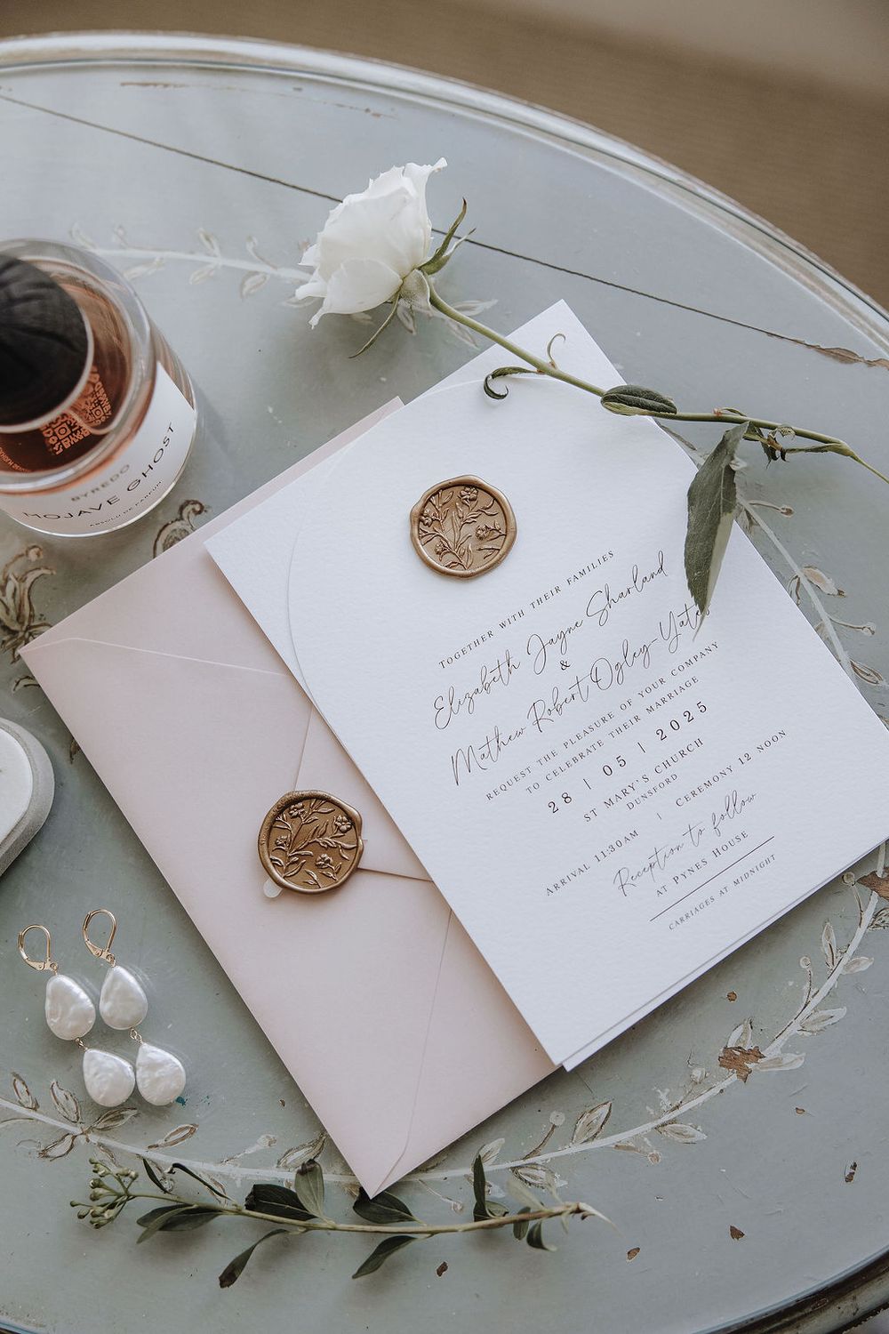 Elegant wedding invitation with gold wax seals and a white rose on a vintage blue-gray table.