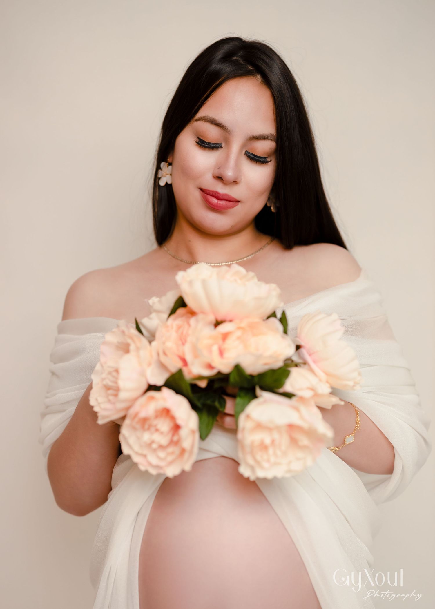 Person wearing off-shoulder white dress holding delicate peach roses in soft studio portrait.