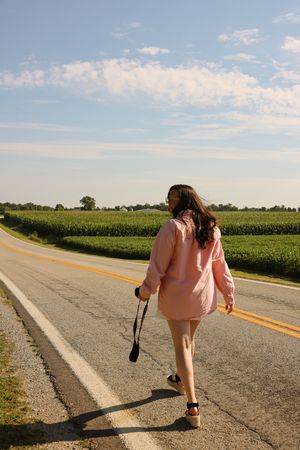 A person in pink walks down a rural road past cornfields under a bright summer sky.