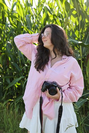 A photographer in a pink shirt and white skirt poses with her camera in a cornfield.