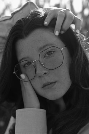 A black and white portrait of a young woman wearing round glasses and touching her hair.