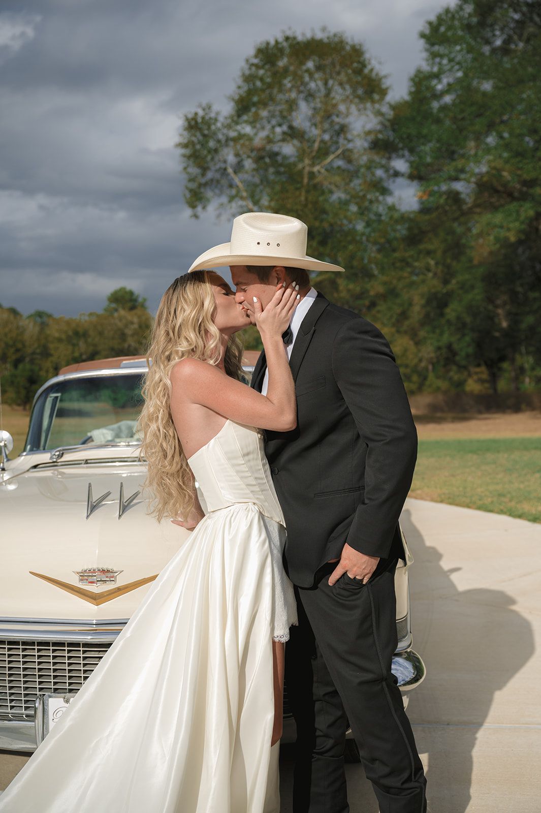 Romantic wedding scene with a couple sharing a kiss beside a classic white Cadillac on an overcast day.