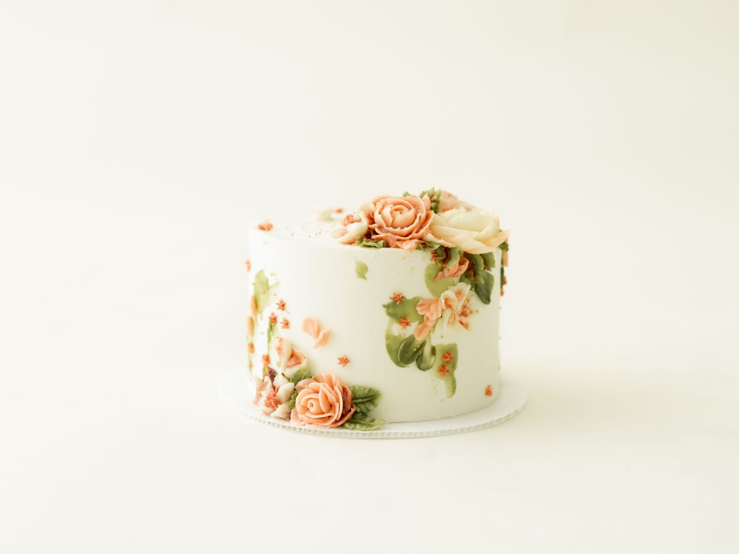 A white buttercream cake decorated with delicate peach-colored roses and green foliage against a light neutral background.