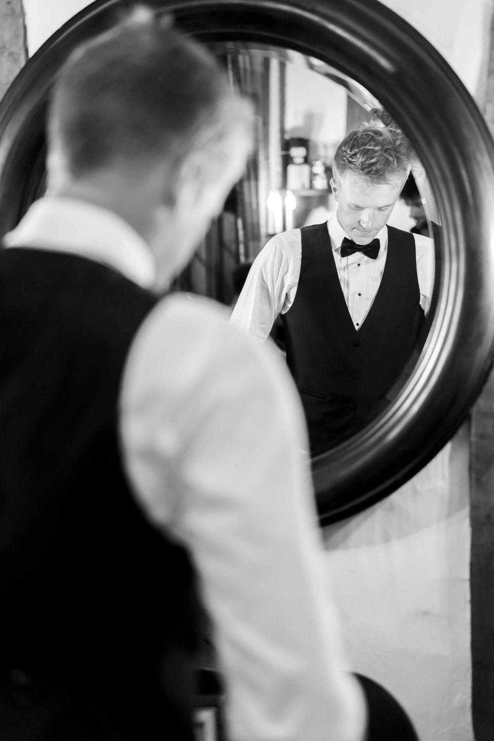 A black and white reflection in a round mirror shows someone adjusting their bow tie while wearing formal attire.
