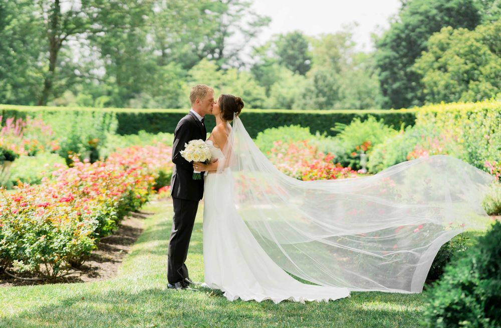 A couple shares a romantic moment in a blooming garden with the bride's veil flowing gracefully in the breeze.