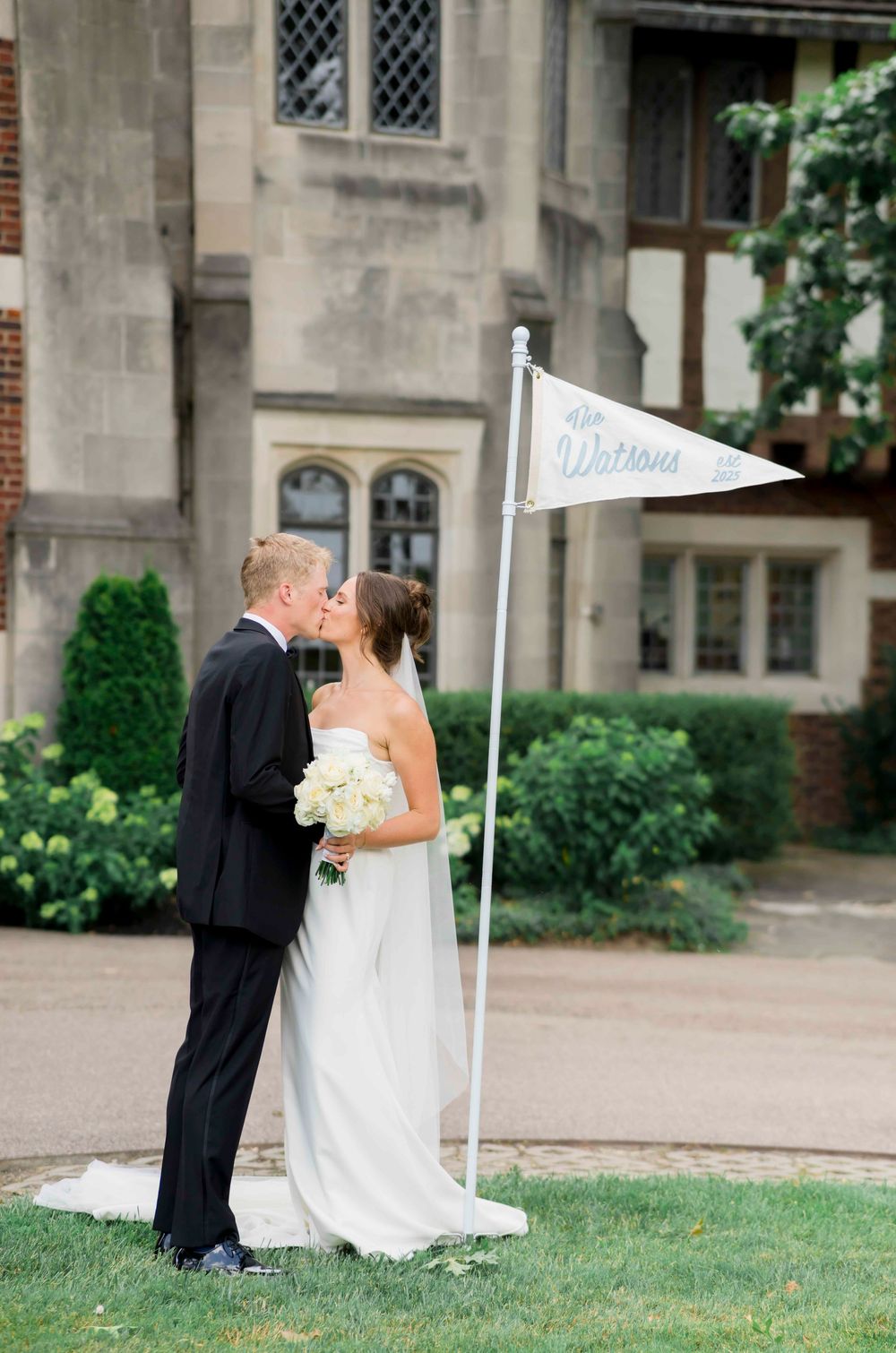 Newlywed couple shares a romantic kiss on a manicured lawn outside a stone Gothic church with white pennant flags.