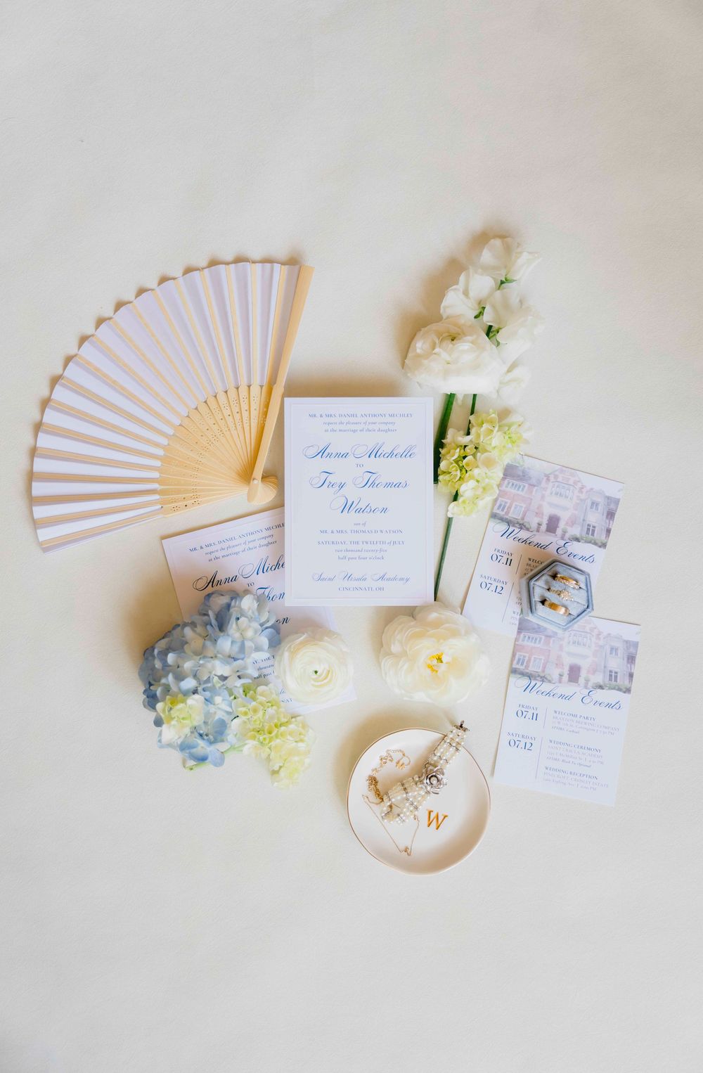 Elegant wedding details flatlay with paper fan, blue hydrangeas, invitation suite and delicate white florals on light background.