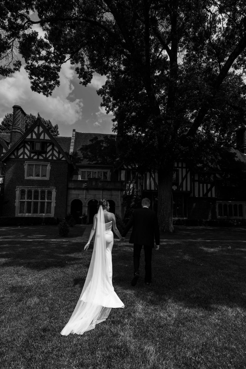 A black and white wedding photo showing a long flowing bridal veil against the backdrop of a Tudor-style mansion.