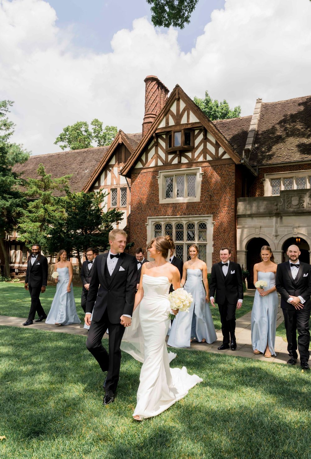 A wedding party walks on a lush lawn in front of a Tudor-style mansion with bridesmaids in light blue dresses.
