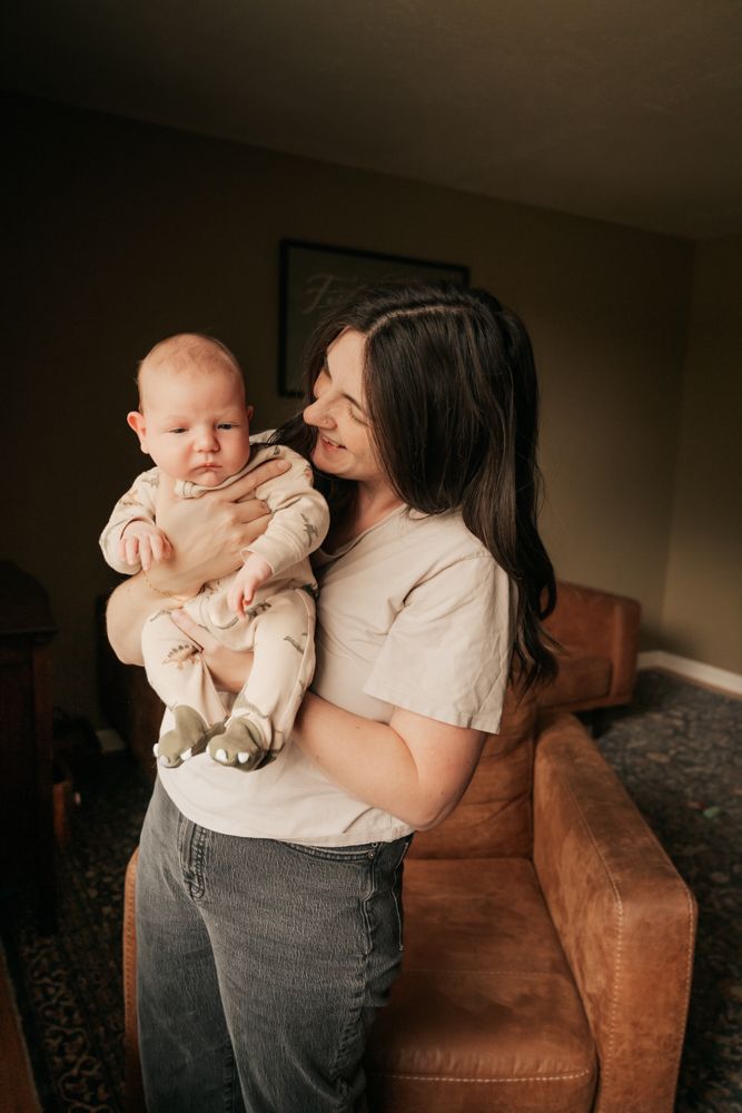A tender moment between mother and baby in front of a brown leather couch in warm indoor lighting.