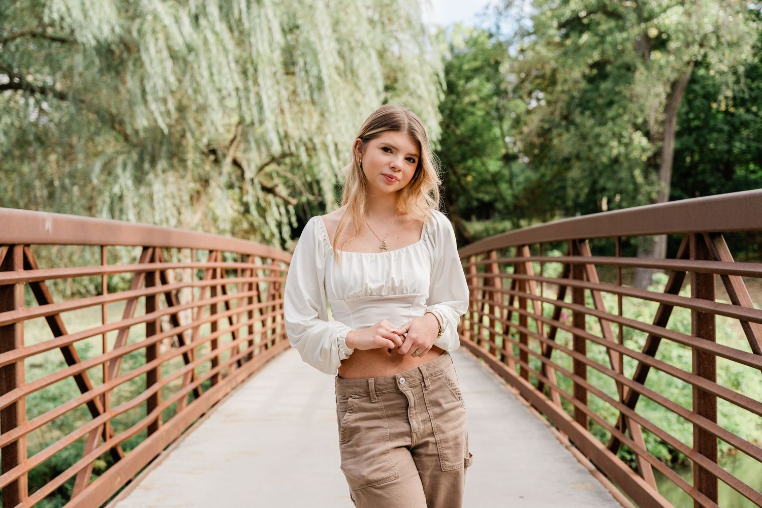 Perrysburg High School senior in a white blouse and khaki pants poses on a bridge surrounded by lush willow trees for photo session.