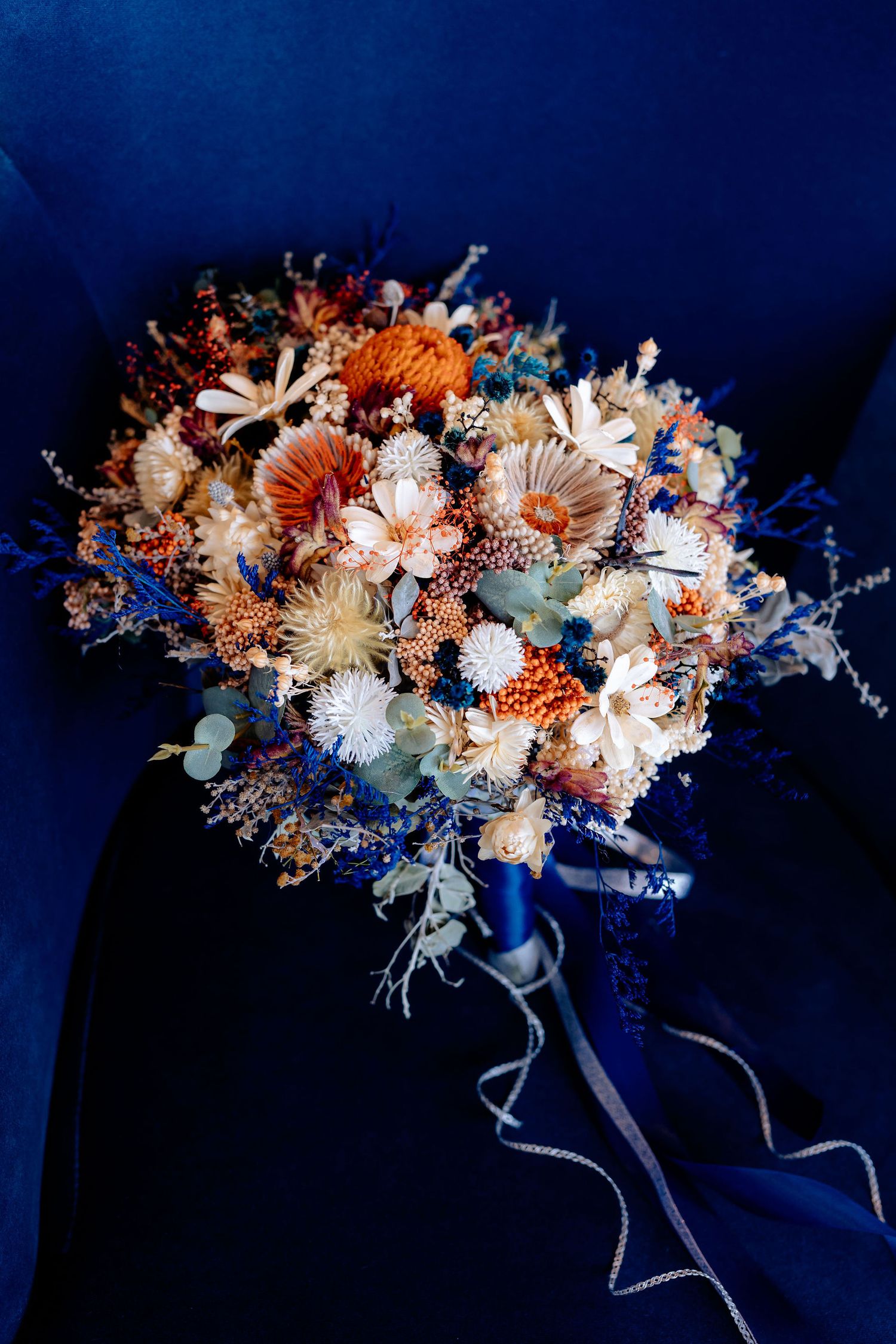 A vibrant dried flower wedding bouquet with orange and white blooms against a navy blue fabric background.