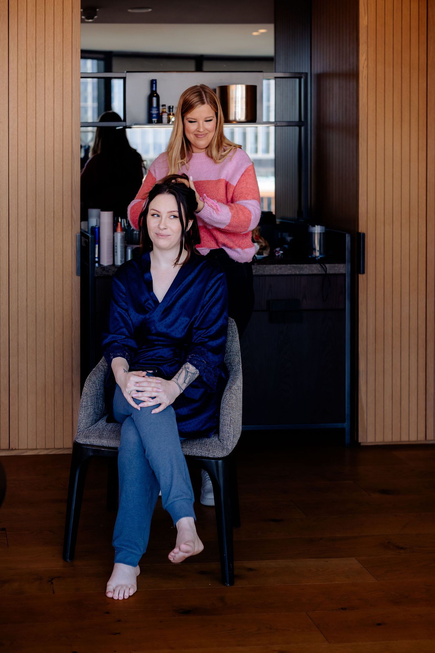 Two people in a modern interior space with wooden paneling, one seated in a gray chair wearing blue while another styles their hair.
