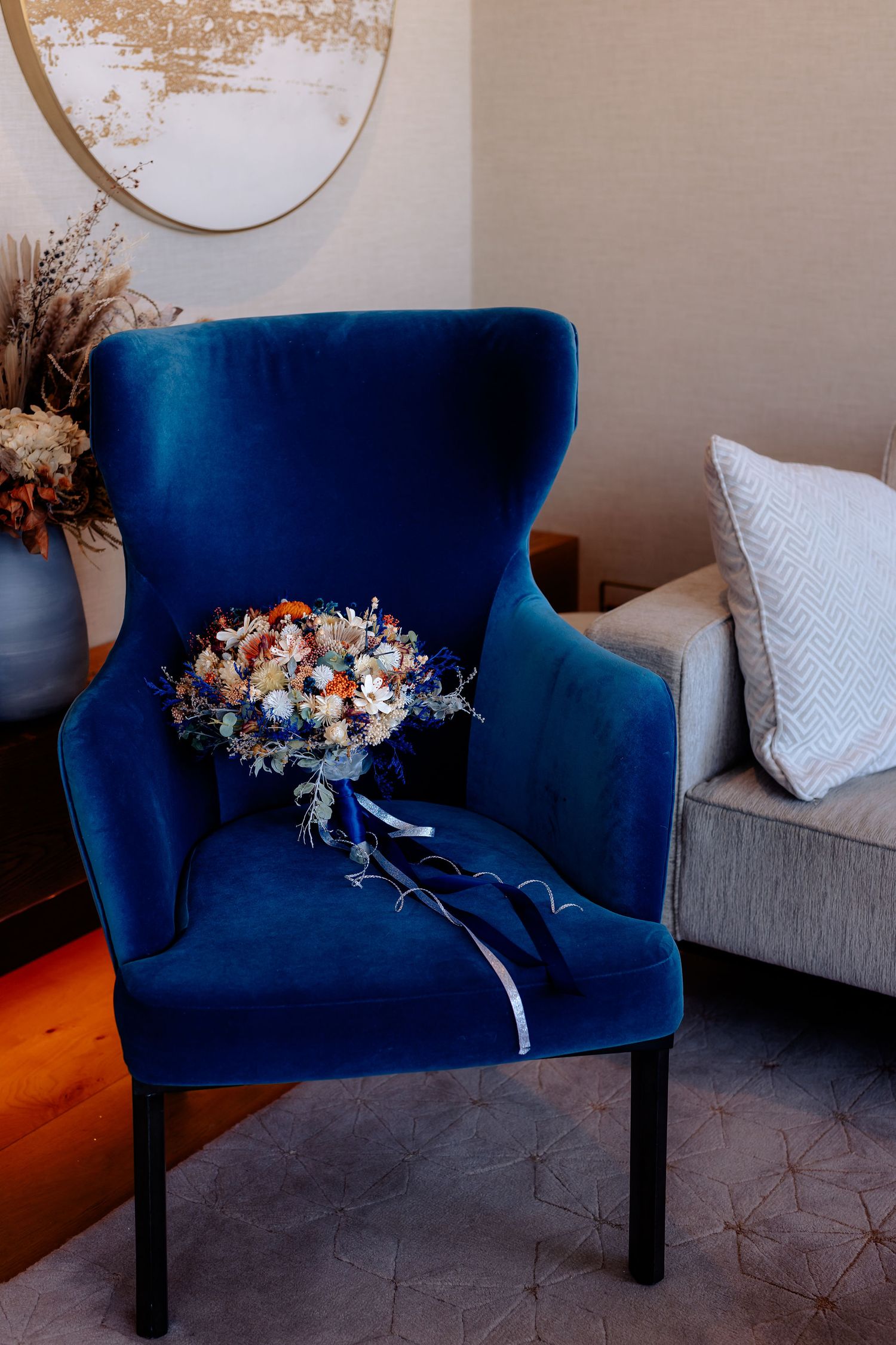 A vibrant blue velvet armchair with a dried floral bouquet resting on its seat near a round mirror and gray couch.