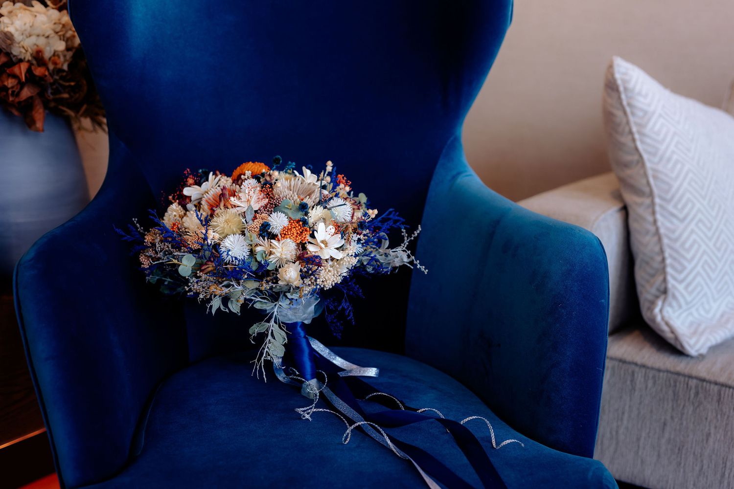 A vibrant wedding bouquet with orange and white flowers rests on a royal blue velvet armchair.
