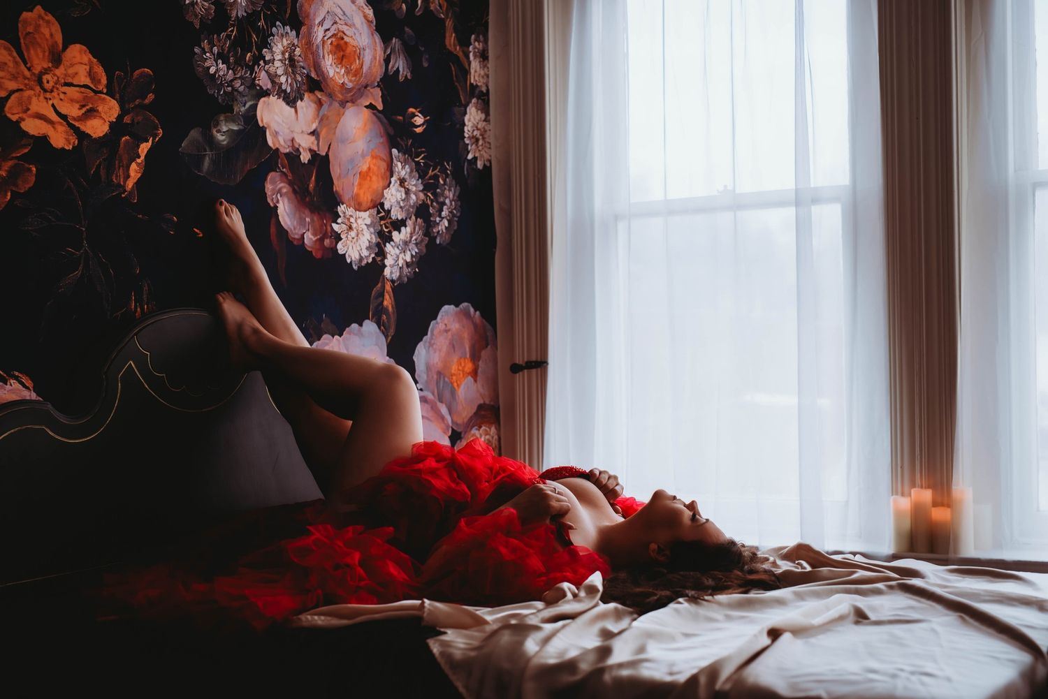 Moody bedroom scene with dark floral wallpaper, red roses scattered on white bedding, and soft window light.