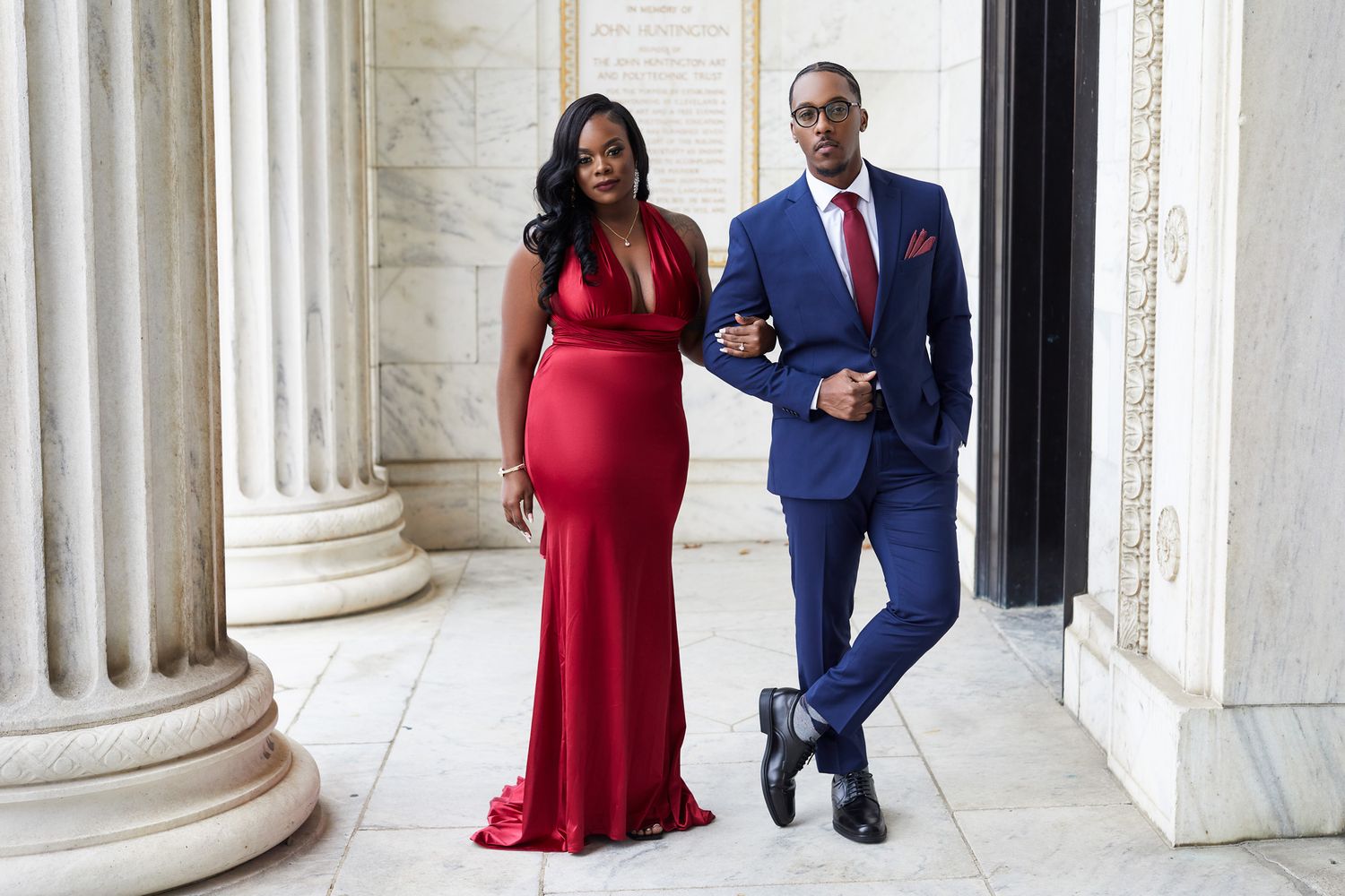 Formal portrait of a couple in evening wear - red gown and blue suit - posed by classical columns.