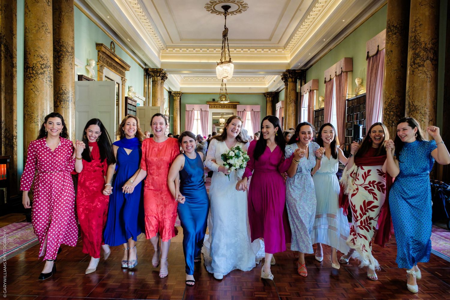 A group of bridesmaids in colorful dresses pose together in an elegant hallway with ornate columns and chandeliers.
