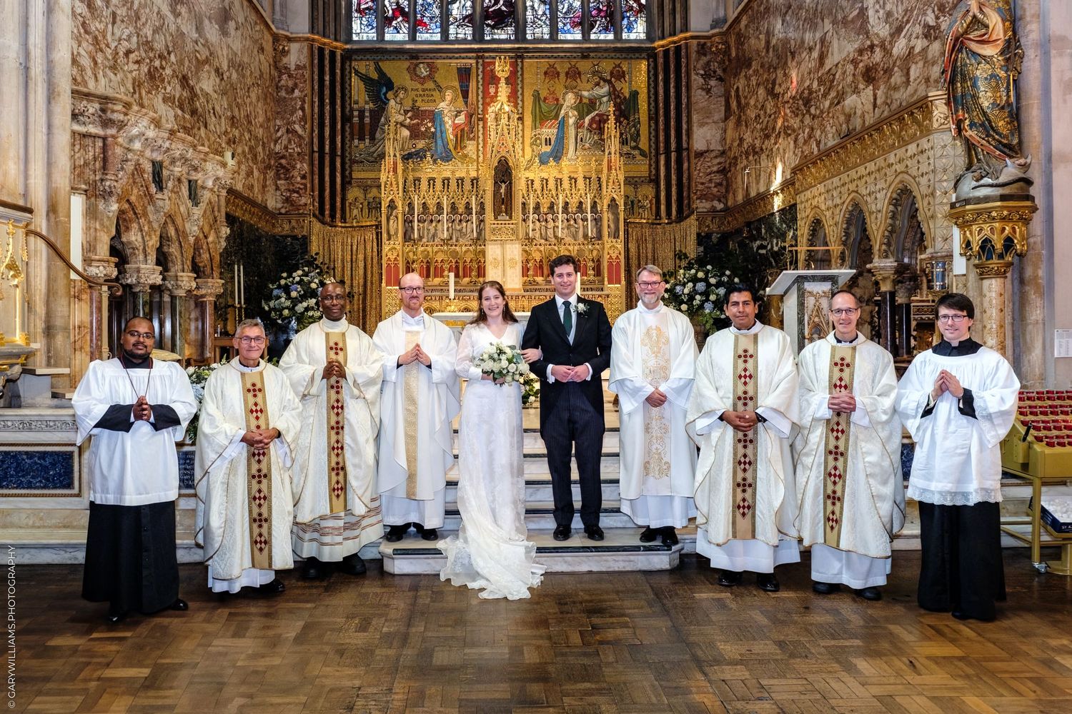 Religious ceremony at ornate church altar with clergy in white and gold vestments and altar servers holding candles.