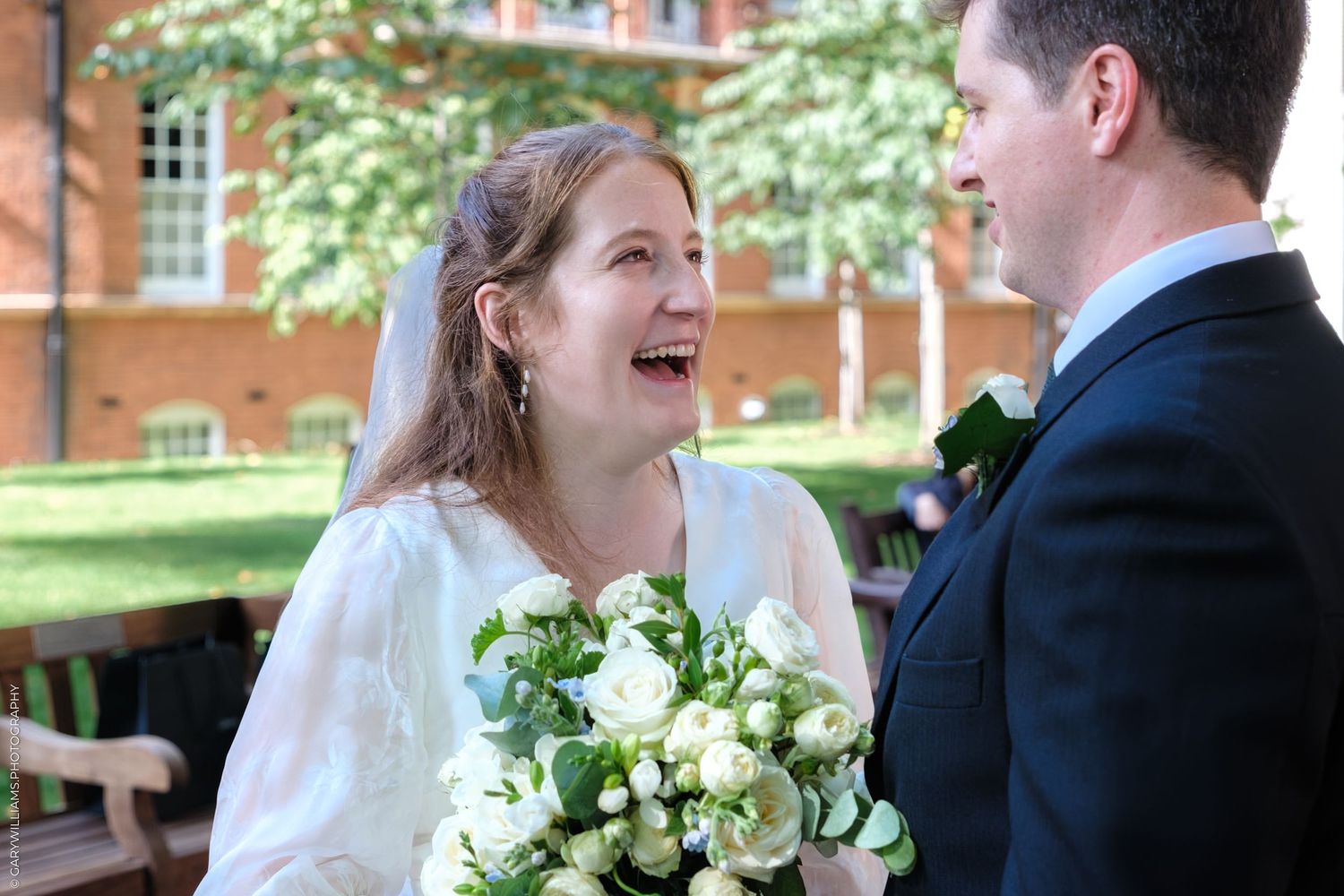 Joyful couple shares a candid moment together at their outdoor wedding ceremony, with white roses and greenery bouquet.