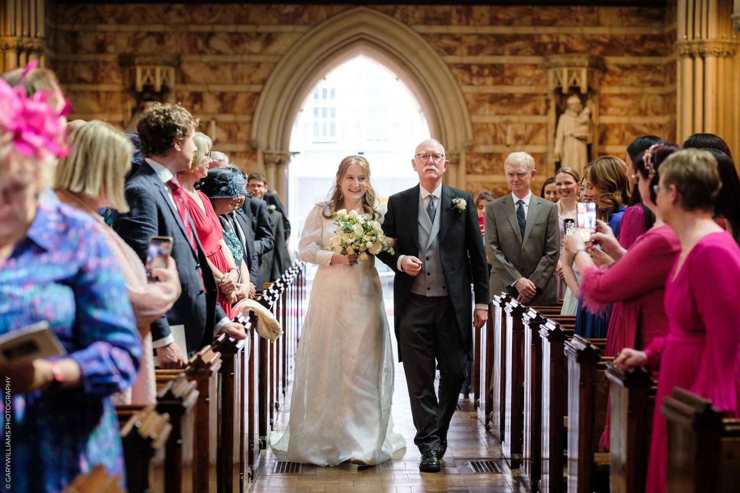 Two people walking down a church aisle lined with guests while carrying a bouquet during a traditional wedding ceremony.