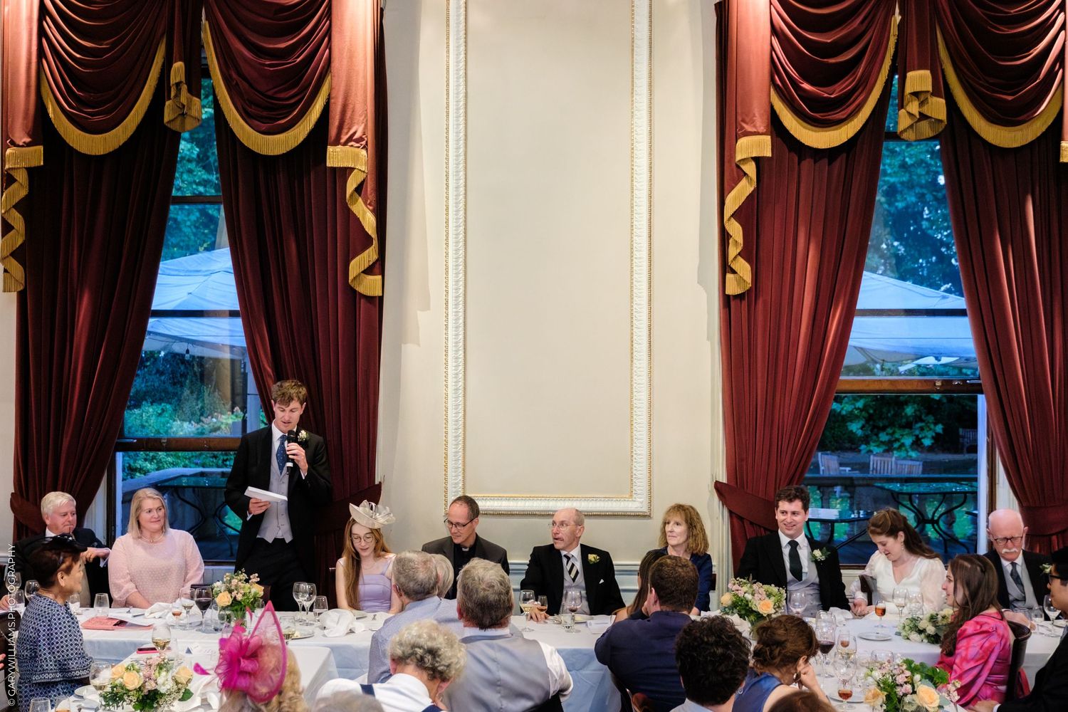 A formal dinner reception in an elegant ballroom with red curtains and guests seated at round tables.