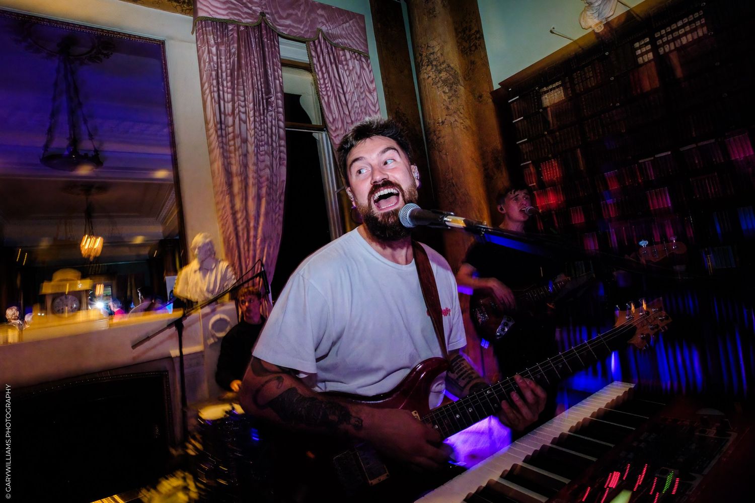 A performer sings while playing keyboard at a dimly lit venue with purple lighting and curtains in the background.