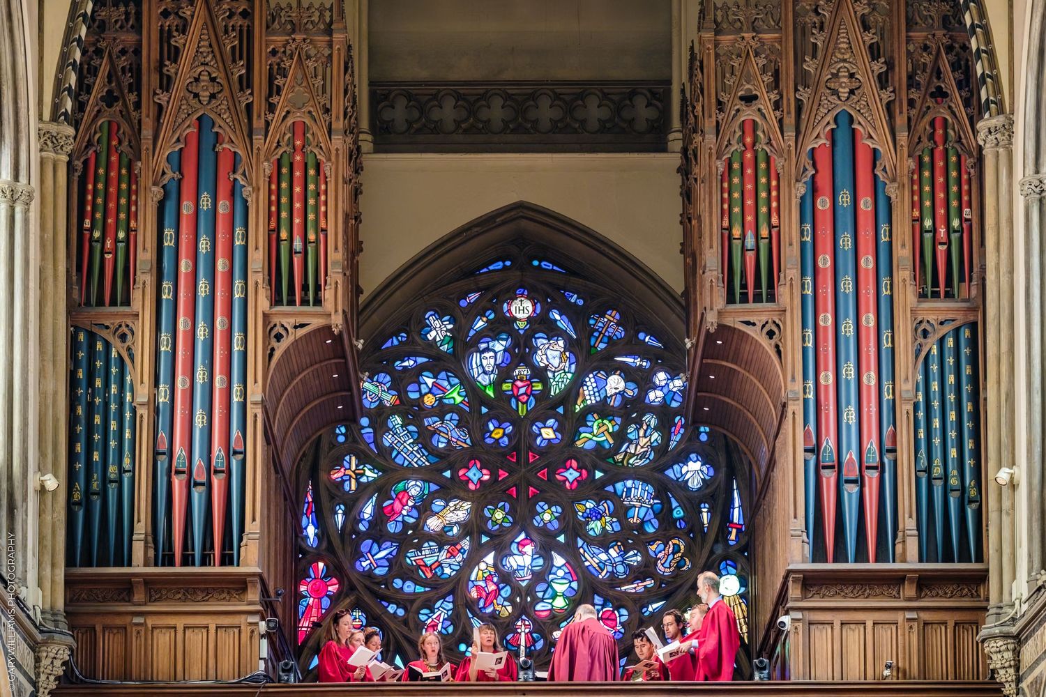 A grand church organ with ornate wooden carvings and colorful stained glass windows sits in a cathedral interior.
