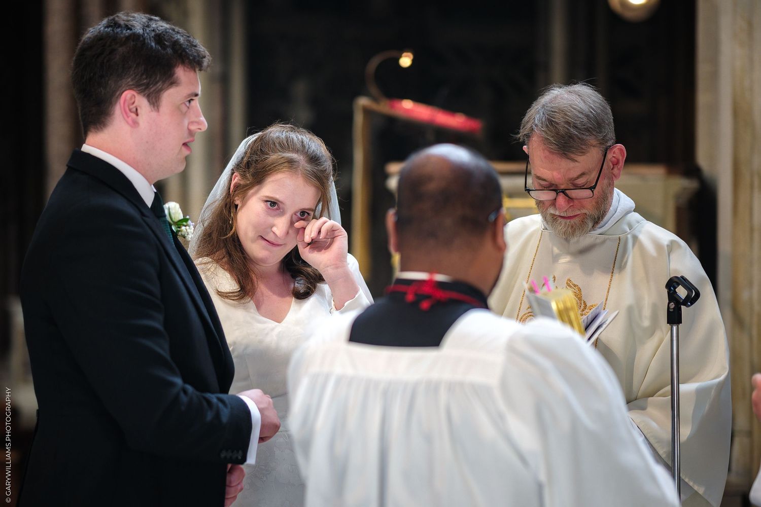 A bride wearing a white dress and her groom stand at the altar with clergy members during a traditional wedding ceremony.