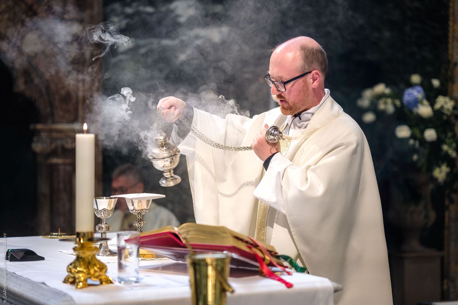 Priest in white robes performs Catholic mass ceremony with incense smoke rising from thurible at church altar during service.