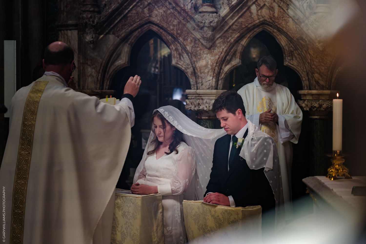 A couple kneels at a church altar during their wedding ceremony while a priest performs the blessing ritual.
