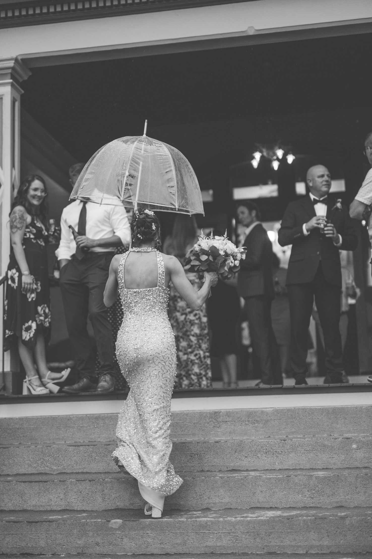 Back view of a bride in a fitted lace dress holding a clear umbrella while walking among wedding guests at an event.